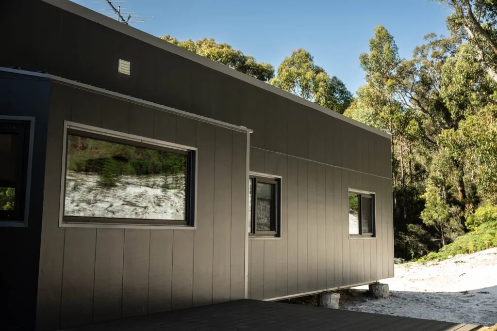 An exterior photo of a grey modular home with three windows and surrounded by trees
